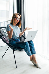 Portrait of a shocked young woman using laptop while sitting in armchair at home