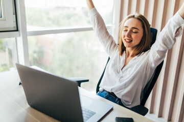 Young attractive woman at a modern office desk, with laptop, stretching her arms with extreme joy...