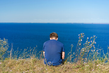 A blond man in a blue shirt sits on a cliff overlooking the blue sea and Islands