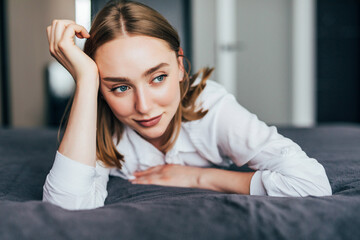 Fototapeta premium Young woman lying at the end of the bed underneath the quilt and smiling, with her head resting upon her hand with the other in her hair.