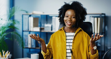 Portrait of pretty cheerful African American woman sitting at office and talking in headset. Beautiful joyful female worker speaking and explaining at working place indoor. Profession concept