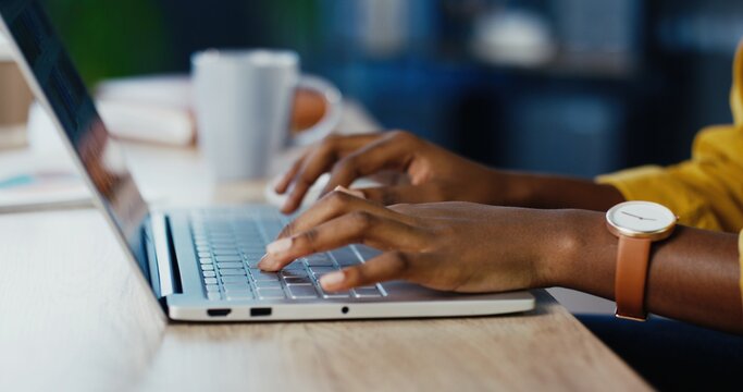 Close up shot of African American female hands typing on laptop while sitting at office desk indoors. Woman fingers tapping and texting on computer keyboard while working in cabinet. Work concept