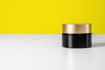Closed jar with gold lid for cosmetics stands on table on yellow background, front view, copy space. Skin care products with moisturizing, nourishing or anti-aging effect.