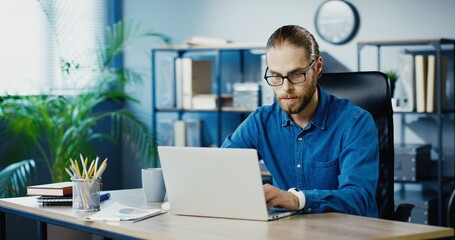 Caucasian serious male office employee typing on laptop while working in cabinet. Handsome busy man in glasses browsing on computer while sitting at workplace indoor. Business concept