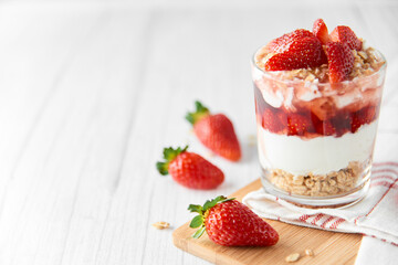 Homemade layered dessert with fresh strawberries, cream cheese or yogurt, granola and strawberry jam in glass on white wood background. Healthy organic breakfast or snack concept. Selective focus.