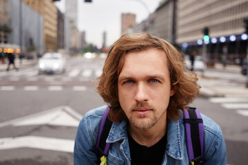 Beautiful long hair young casual wear tourist man with black t-shirt and jeans jacket sitting on metal fence looking straight to camera with big city background. Travel and lifestyle concept.