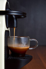 Espresso machine making coffee with capsules in a glass cup on wooden table. Front view. Copy space. Selective focus..