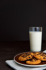 Tasty Chocolate chip cookies on the brown plate and stripped napkin and glass of milk on dark background. Food, junk-food, culinary, baking and eating concept. Selective focus. Copy space.