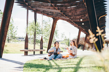 Young lady in denim suit shoots positive girl friend playing ukulele with modern smartphone spending time in contemporary city park on sunny day.