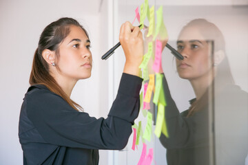 Young Latin businesswoman writing on sticker with marker. Focused confident beautiful brunette...