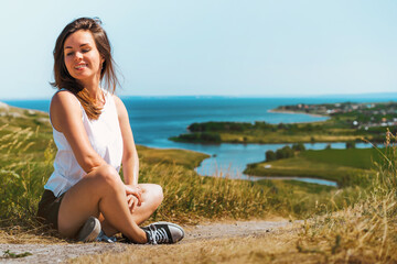 The girl is sitting cross legged on the ground against the background of a mountain landscape with a river and fields and meadows