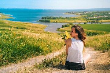 A young girl sits with her back to the camera on a cliff holding a sunflower in her hand with a view of fields and Islands in the sea, green meadows