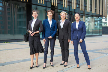 Fototapeta premium Happy female professional team standing together near office building, posing, looking at camera and smiling. Full length, front view. Businesswomen group portrait concept