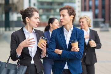 Female business colleagues with takeaway coffee mugs walking together in city, talking, discussing project or chatting. Medium shot. Work break concept