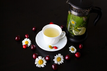 A white Cup with herbal tea, next to a teapot that brews currant leaf, mint, chamomile and lemon. On a black background with daisies and cherries.