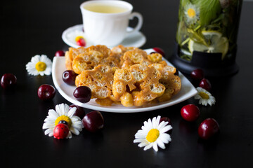 On a white plate is a dessert of toffee and corn sticks,decorated with daisies with ladybirds and cherries.Next to it is a Cup of herbal tea and a teapot in which lemon ,mint and chamomile are brewed.