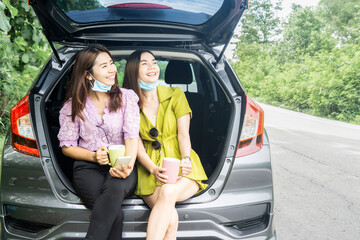 two happy Asian woman wearing protective mask sitting on hatchback car hand holding coffee cup travel with new normal lifestyle 