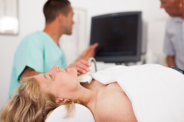 Skilled doctor doing ultrasound diagnostics of thyroid to woman lying on couch in medical office, her husband on background