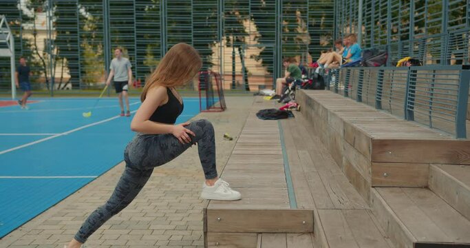 Sport girl is doing a stretch. She is leaning on the step. Her hair is flying in the wind. She's wearing a sports uniform. The camera is moving from left to right. People are playing