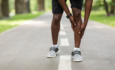 Black sportsman touching his aching leg while jogging outdoors