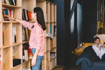 Young woman in casual outfit standing near shelves choosing book for read enjoying literature on free time, intelligent female student choosing text book for learning spending time in library.