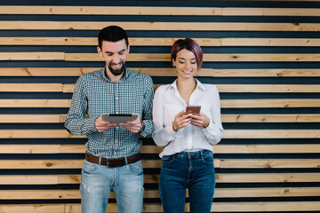 Cheerful young man and woman standing against wall and chatting online in social networks on smartphone and tablet.Young people from millennial generation using internet for communication