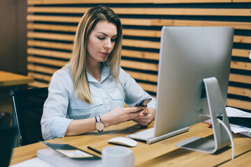 Pensive young woman checking email and reading notification on smartphone connected to 4G internet during work at modern computer.Female office employee updating profile on phone sitting on workplace