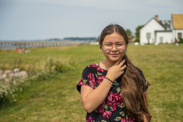 A young preteen girl with long brown hair looks straight into the camera and smiles. A blue hazy sky in the background