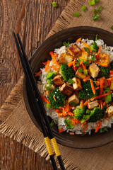 Tofu with rice and vegetables. Served on brown plate with chopsticks. Top view.