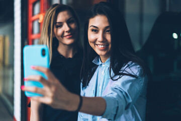 Young charming prosperous hipster girls taking picture together on smartphone camera for updating profile picture in network.Cute smiling women enjoying leisure time in office during work break