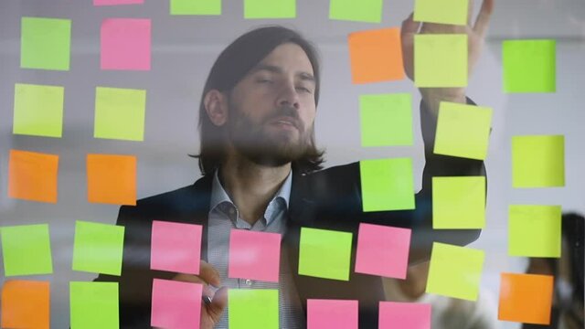 Focused male executive writing business ideas list on post it sticky notes attaching replacing stickers to scrum glass board in office developing work organization manage tasks project strategy plan
