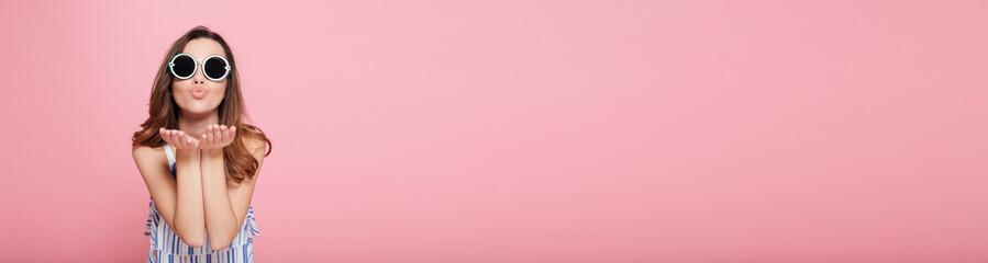 Cute young woman in round sunglasses standing and sending kiss