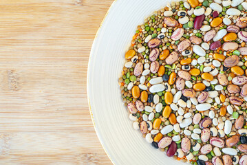 Pile of legumes including beans, peas, lentils arranged on a white plate placed on a wooden cutting board.