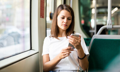 Portrait of modern female passenger in T-shirt using mobile phone in tram