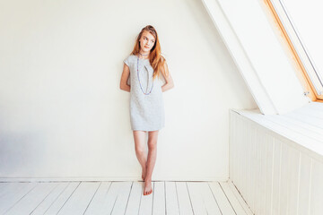Fototapeta premium Happy teenage girl smiling. Portrait young happy positive teen woman in grey dress at home in bright room against white wall. European woman. Positive human emotion facial expression body language.