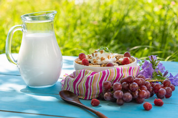Breakfast cereals with milk and raspberries, grapes, banana in the open air on a light blue table and on a background of green grass.