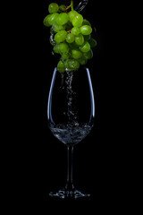 Wine glass on black background with  bunch of green grapes on top of it and white wine or liquid flowing on it