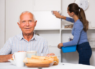 Portrait of elderly man at tea table, young woman cleaning furniture at home kitchen