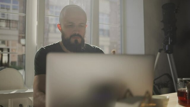 Freelancer programmer working from home on kitchen and using laptop. Bearded man working with a computer, prints text on keyboard. Self entrepreneur sitting and working at his modern apartment.
