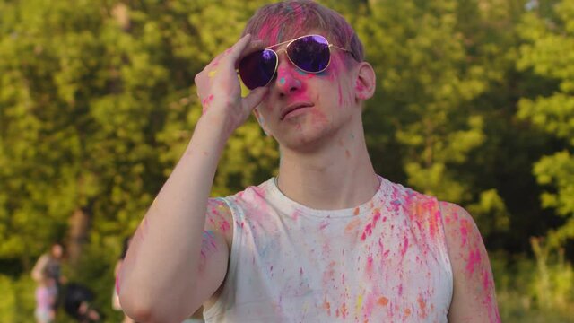 Dirty guy in colored paints, smiling looks at the camera and takes off his sunglasses. Cool guy in colored powder takes off his glasses and looks at the camera