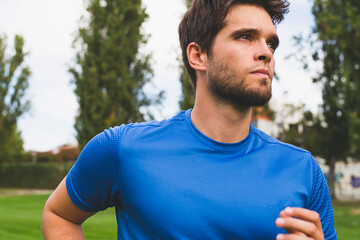 Close-up of attractive young man with a beard and blue t-shirt runs through the park in good weather.