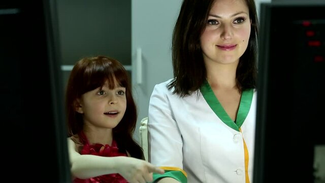 Professional attractive young female doctor smiling showing computer screen monitor to little cute happy girl in office