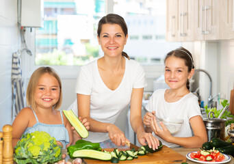Naklejka premium Happy mother with two little daughter cooking vegetable salad at home kitchen