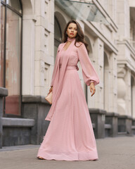 Fashion outdoor portrait. Young stylish woman in long pink evening dress walking at city street on...
