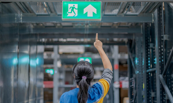 The back view woman pointing at the emergency fire exit sign show the way to escape at the public area, warehouse store.