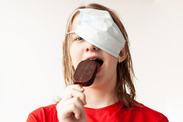 A teenage girl with blonde hair pushed a medical mask up on her hair, bites off a chocolate ice...