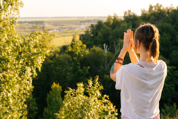 Fototapeta premium Relaxed young woman standing in Namaste pose, nature outside in evening at sunset background of beautiful nature, landscape and scenery. Happy girl performing meditating, bright rays of evening sun.