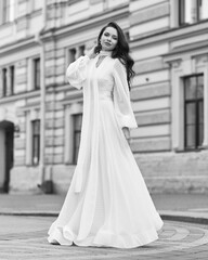Obraz premium Elegant fashionable stylish female model in long white dress walking at city street. Outdoor full length portrait. Caucasian lady with wavuy hair and makeup