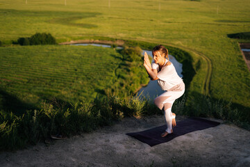 Concentrated woman yoga teacher practicing yoga, standing in Eagle exercise, Garudasana pose on top of cliff background of river below and green grass in evening during sunset. 