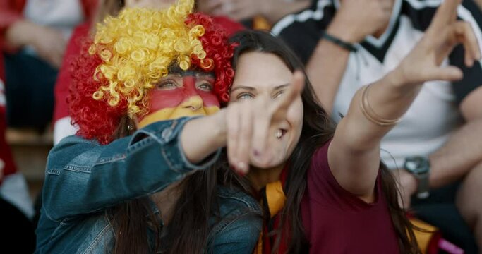 Two women sitting in fan zone with a wig and face painted in german flag colors pointing at a big screen and blowing a kiss. Germany soccer team supporters looking themselves in a big screen at stadiu